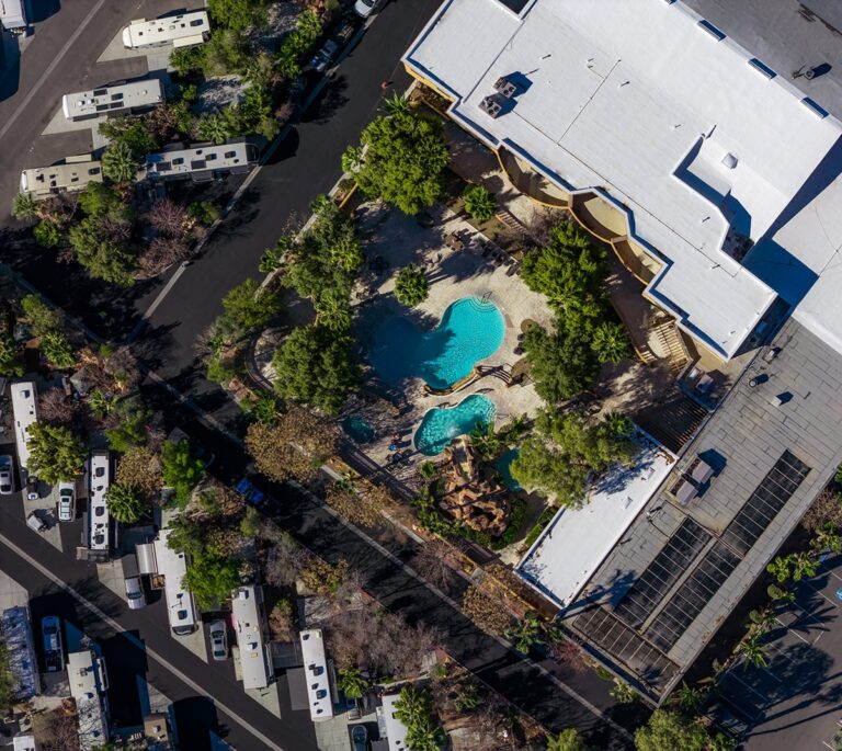 Aerial view of an RV park with a pool area, trees, and parked campers near a large white-roofed building.