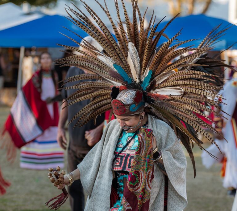 Person in traditional Aztec attire with a large feathered headdress dancing outdoors at a cultural event.