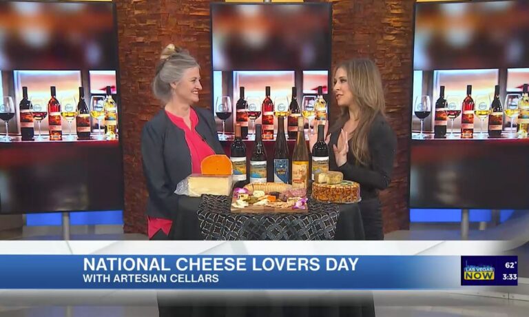 Two women discuss cheese and wine on TV for National Cheese Lovers Day, with cheese and wine bottles on a table.