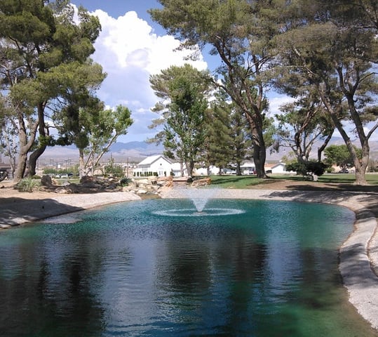 A small pond with a central fountain surrounded by trees and grass, with a house and mountains in the background.