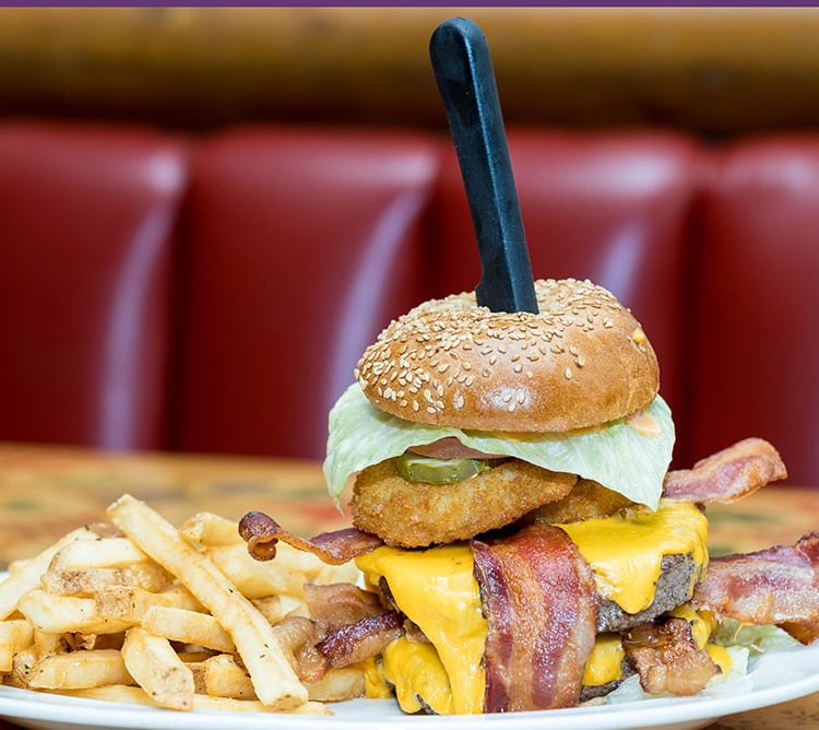 A cheeseburger with bacon, lettuce, onion ring, and a knife in the bun, served with French fries.