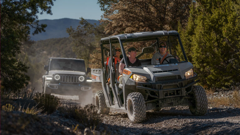 A group rides an off-road utility vehicle on a rocky trail with a Jeep following behind in a forested area.