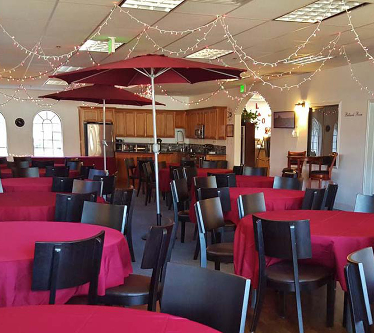 Tables with red tablecloths, black chairs, and large umbrellas in a decorated room with string lights.
