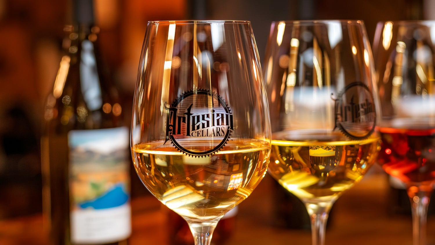 Close-up of Artesian Cellars wine glasses filled with white wine, with wine bottles blurred in the background.