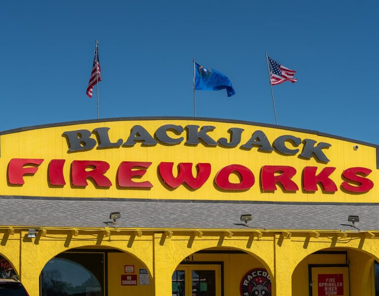 A yellow building with BLACKJACK FIREWORKS sign and three flags on the roof against a clear blue sky.