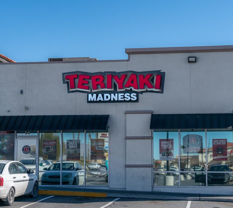 Storefront of Teriyaki Madness restaurant with signs and posters visible on glass windows.