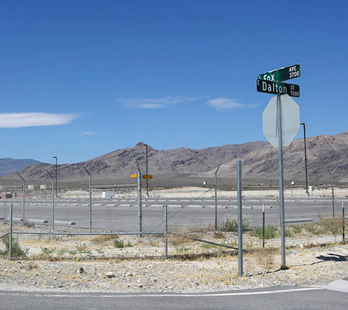 Street signs for Dalton St and Enax Ave near a fenced lot with mountains in the background under a blue sky.