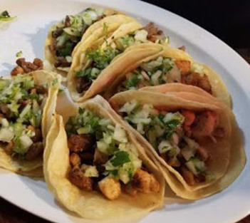 A plate of five tacos filled with diced meat, onions, and cilantro on corn tortillas.