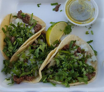 Three street tacos with beef, chopped onions, cilantro, lime wedge, and green salsa in a takeout container.