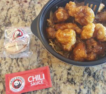A bowl of orange chicken, a fortune cookie, and a packet of chili sauce on a granite countertop.