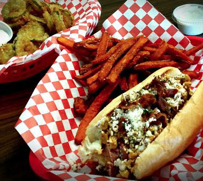 A sandwich with crumbled cheese, sweet potato fries, and a side of fried pickles on checkered paper.