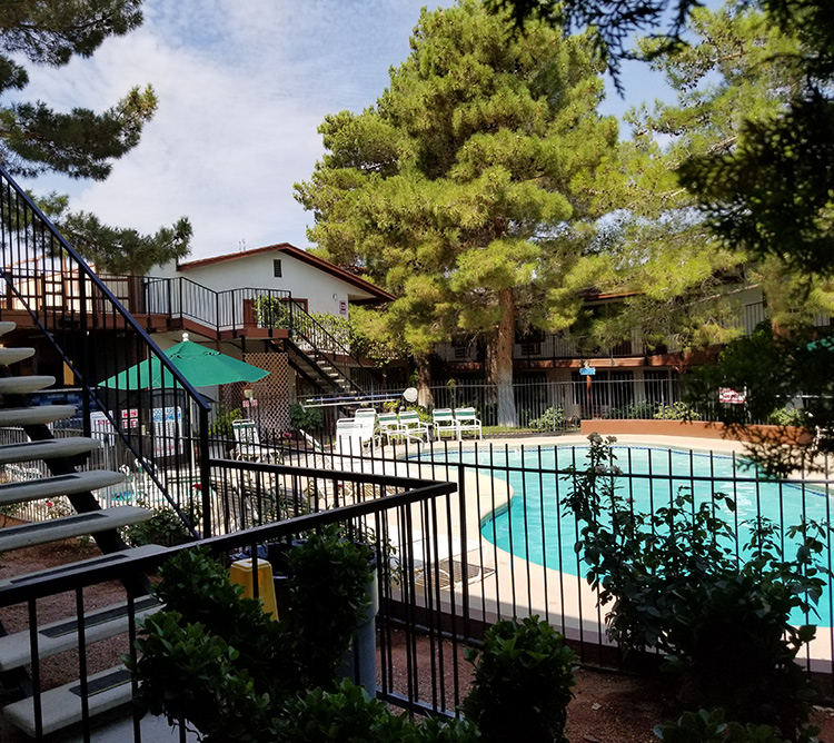 Fenced outdoor swimming pool surrounded by trees, lounge chairs, and a stairway under a partly cloudy sky.