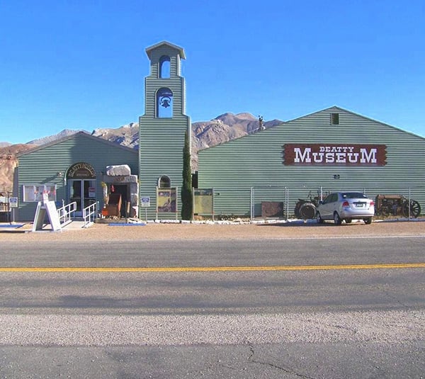 Green building labeled Beatty Museum with mountains in the background and a car parked outside.