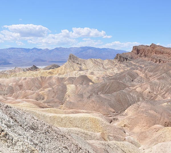 Colorful, rocky badlands under a bright blue sky with distant mountains and scattered white clouds.