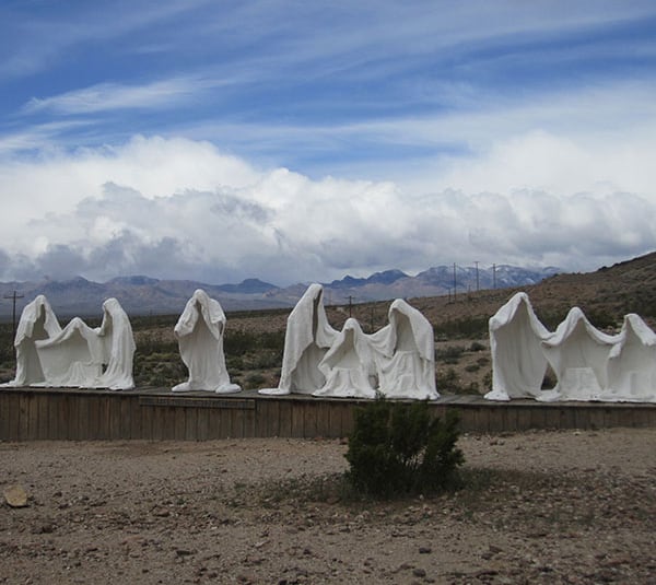 White ghost-like statues stand in a row in a desert landscape beneath a cloudy sky with mountains in the background.