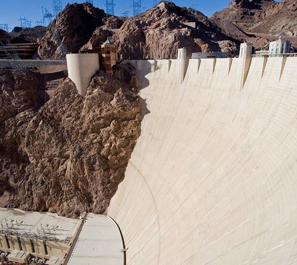 A large concrete dam built between rocky cliffs, with power lines visible in the background under a clear sky.