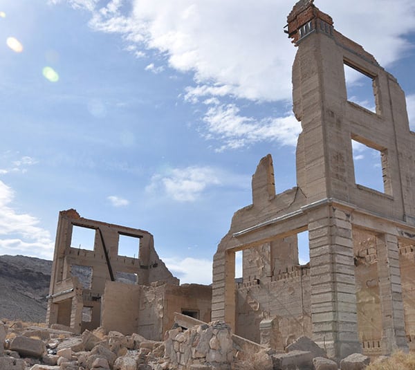 Two crumbling stone buildings stand in a rocky desert under a blue sky with wispy clouds.