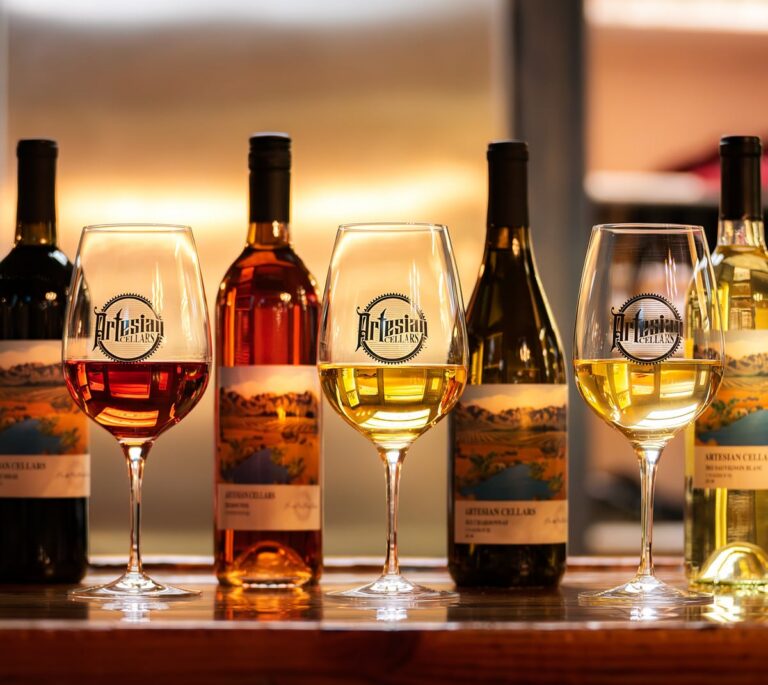 Three glasses of wine and five bottles on a wooden bar, all labeled Artisan Cellars.
