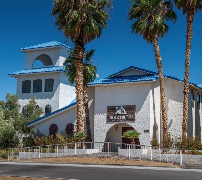 White stucco building with blue roof, palm trees, and a sign reading Charleston Peak Hotel out front.