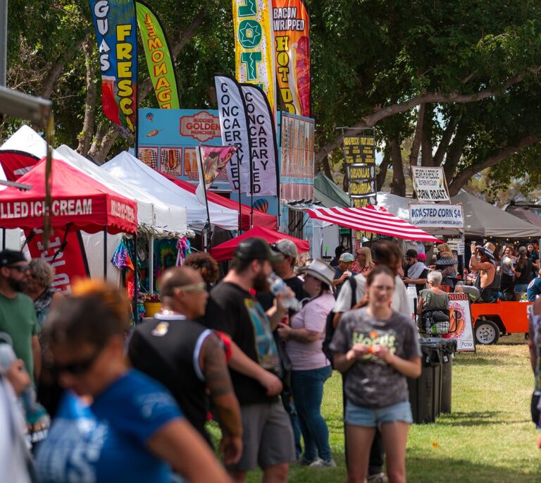 People walk along food stalls with colorful banners at an outdoor fair on a sunny day.