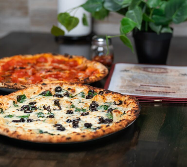 Two pizzas, one with olives and spinach, one with pepperoni, on a table next to a menu and a potted plant.