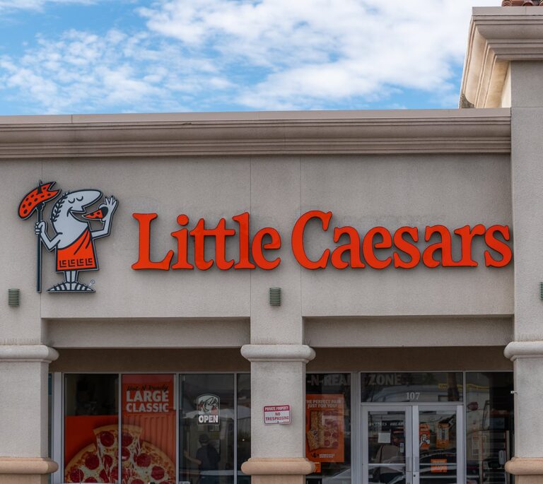Little Caesars storefront with orange sign and mascot holding pizza, under a partly cloudy sky.