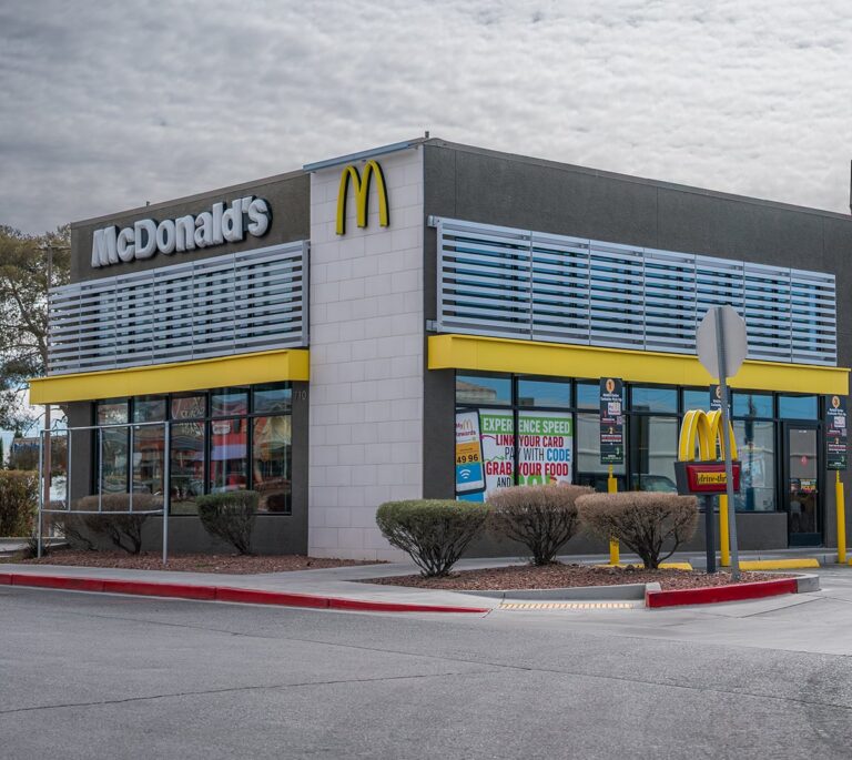 A McDonalds fast food restaurant with drive-thru under a cloudy sky, surrounded by bushes and a street.