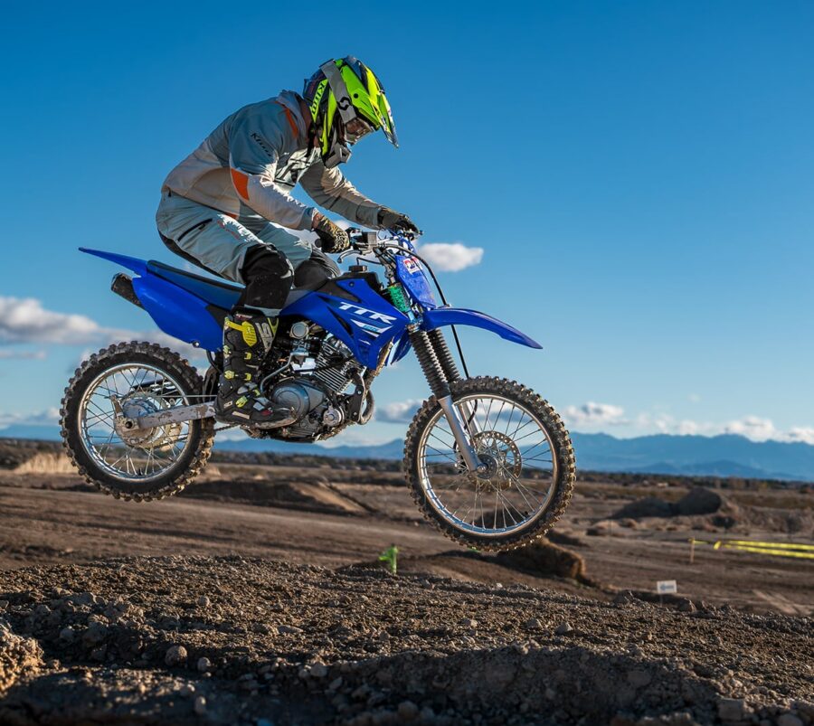Person in motocross gear riding a blue dirt bike, jumping over rough terrain under a clear blue sky.