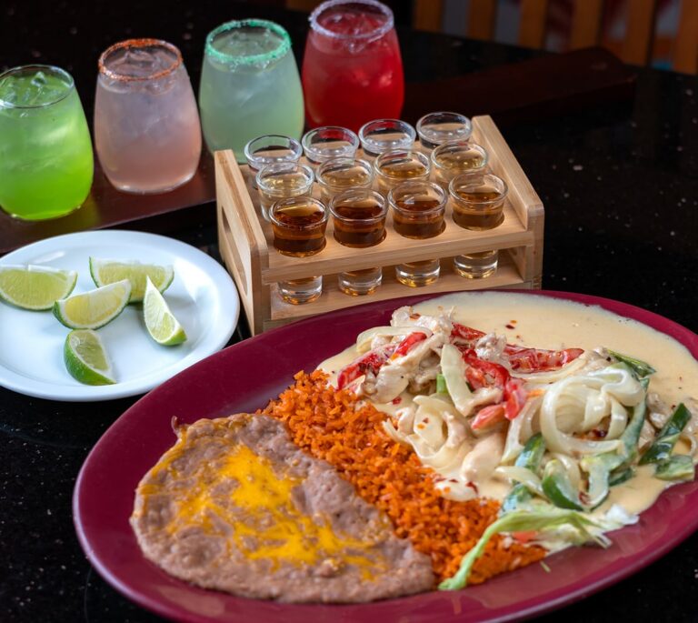 A plate with rice, refried beans, creamy chicken dish, lime wedges, colorful drinks, and a rack of tequila shots.