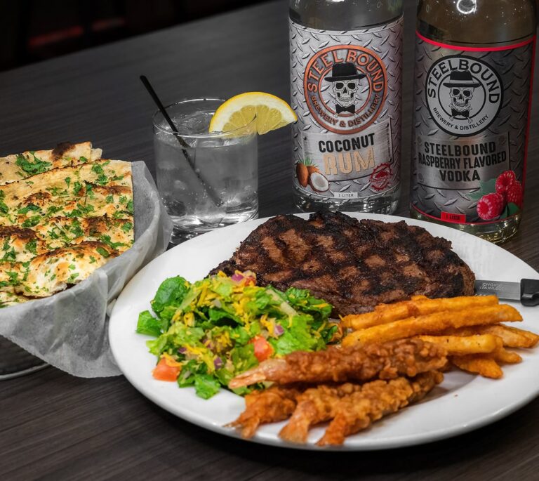Grilled steak, fries, salad, bread, two bottles of spirits, and a drink with lemon on a dark table.
