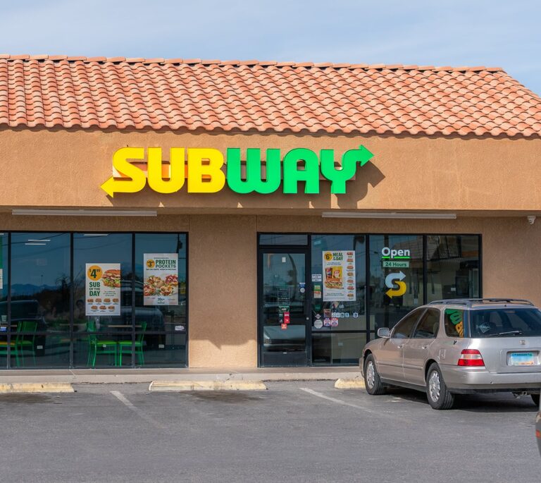 Subway restaurant exterior with large yellow and green sign, windows, and a parked silver car in front.