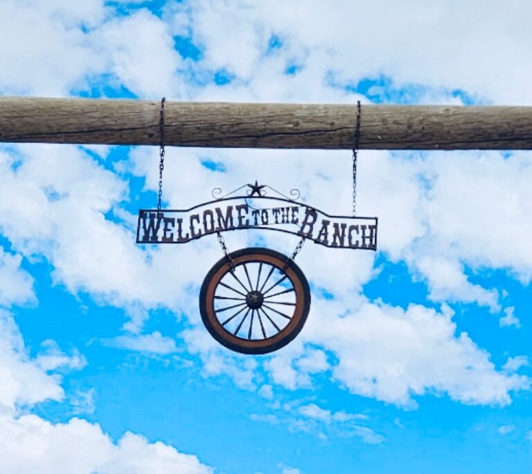 Metal Welcome to the Ranch sign with a wagon wheel hangs on a wooden beam against a blue sky with clouds.