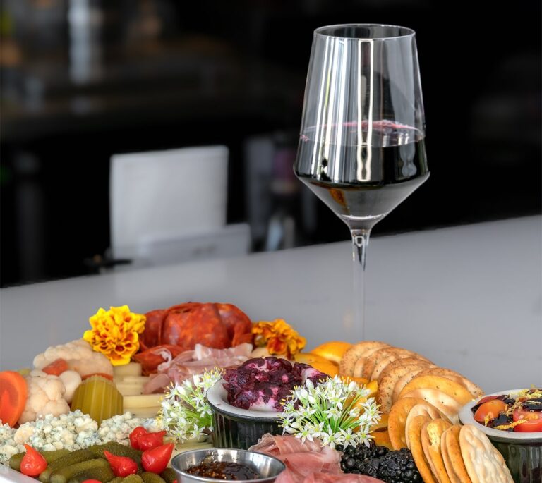 A glass of red wine with a charcuterie board of meats, cheeses, crackers, and garnishes.