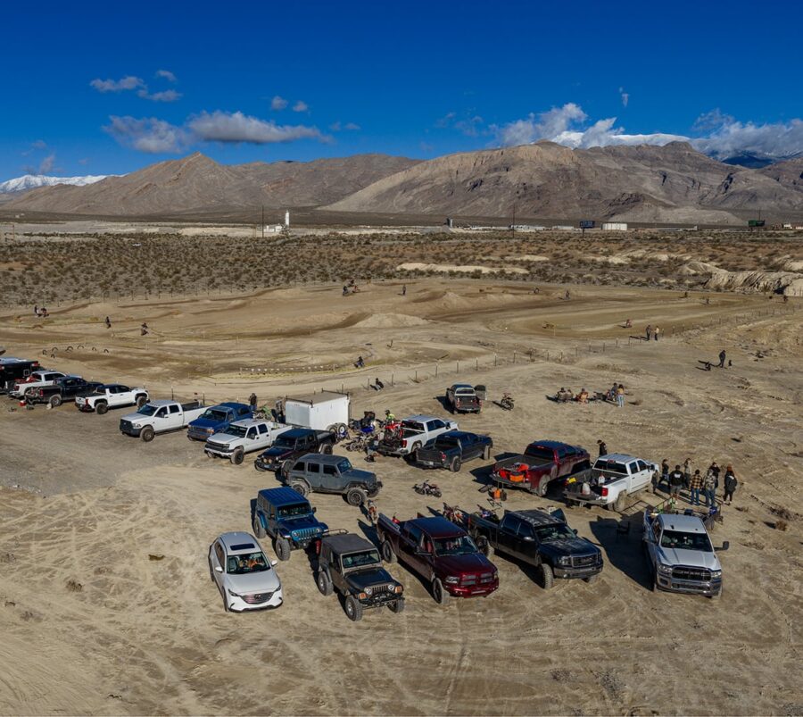 A group of trucks and cars parked on a desert landscape with people gathered and mountains in the background.