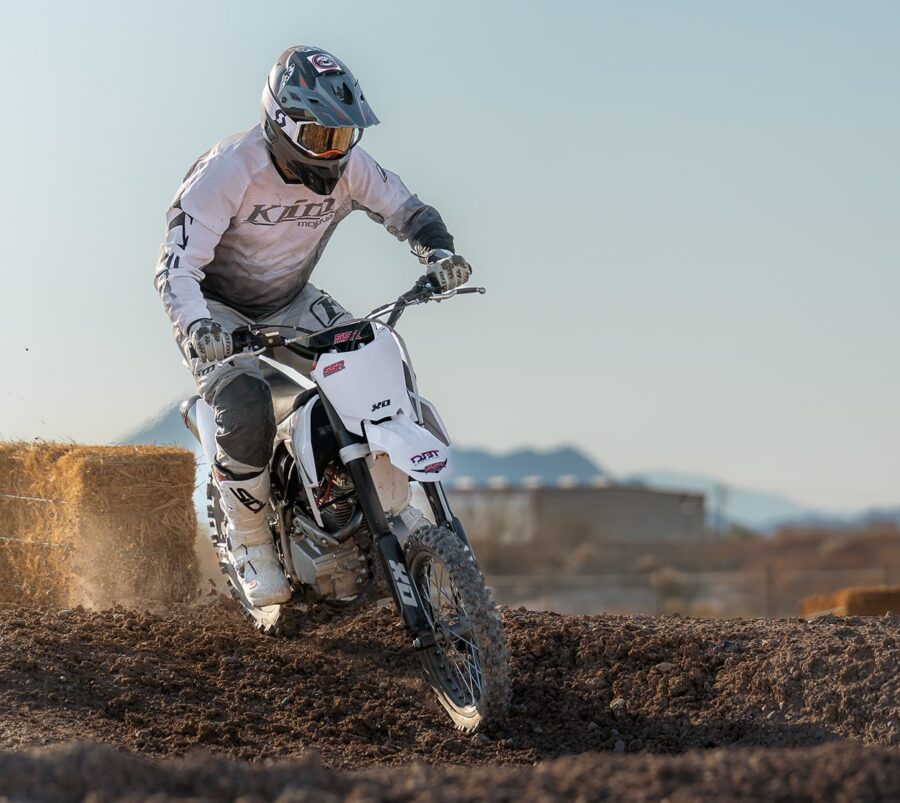 Motocross rider in white gear turns on a dirt track with hay bales and mountains in the background.
