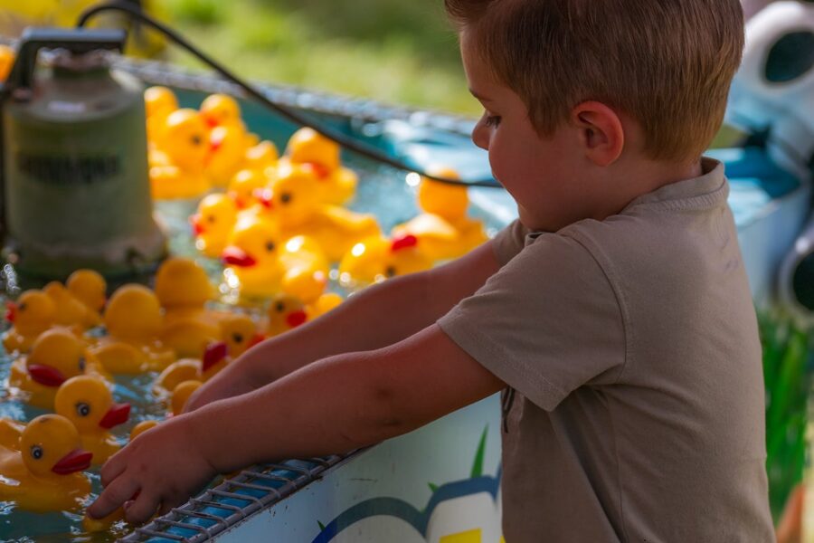 A young boy plays with floating yellow rubber ducks at a water game booth outdoors.