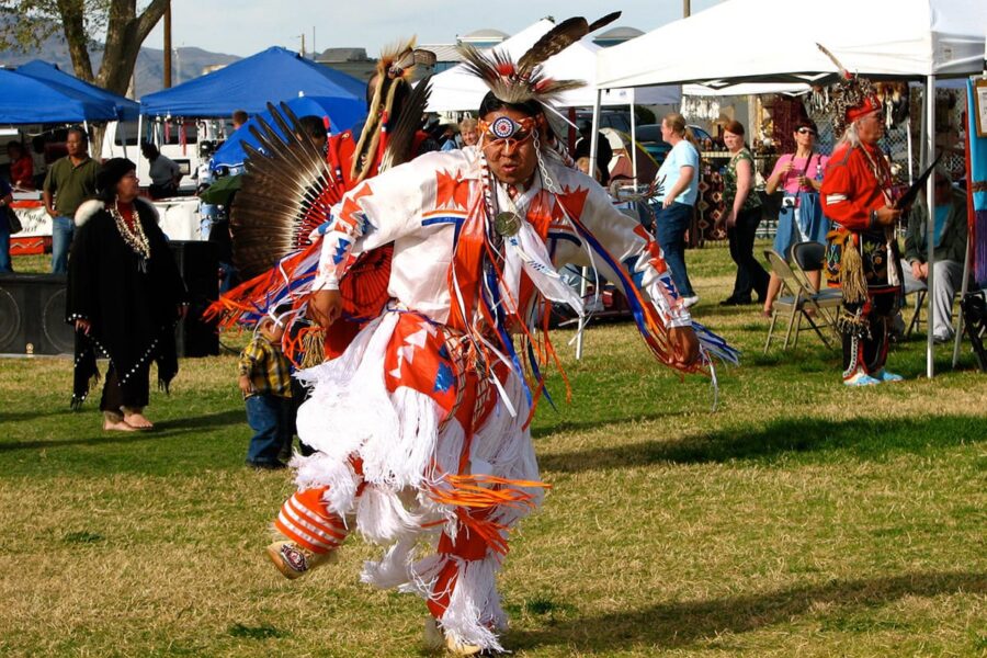 A person in vibrant Native American regalia dances outdoors at a cultural event with tents and onlookers.