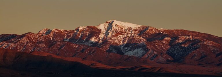 Snow-capped mountain peak at sunset, with reddish light on rugged, rocky slopes and a clear sky.