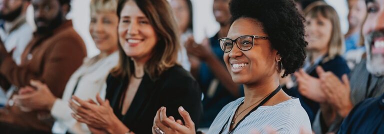 A diverse group of people smiling and clapping while attending an indoor event or presentation.