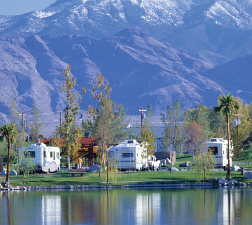 RVs parked by a lake with trees, green grass, and snowy mountains in the background.