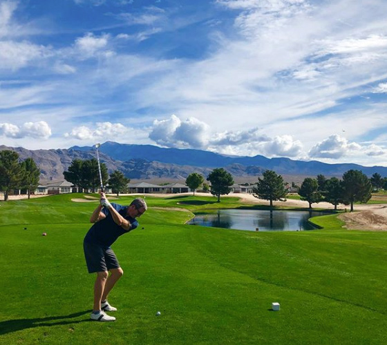 Man preparing to swing a golf club on a sunny golf course with mountains and a pond in the background.