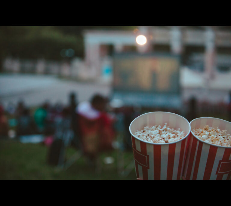 Two striped tubs of popcorn at an outdoor movie night, with a blurry screen and seated crowd in the background.