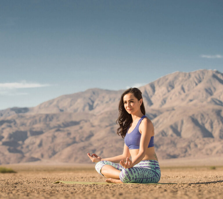 Woman in yoga clothes sits cross-legged meditating on a mat in a desert with mountains in the background.