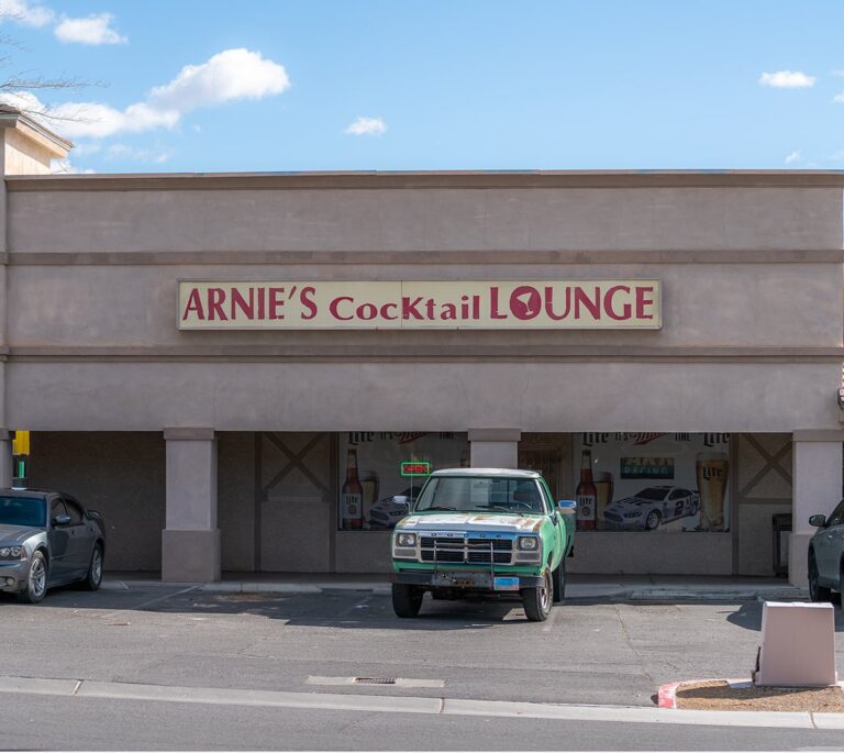 A green truck is parked in front of Arnies Cocktail Lounge on a sunny day.