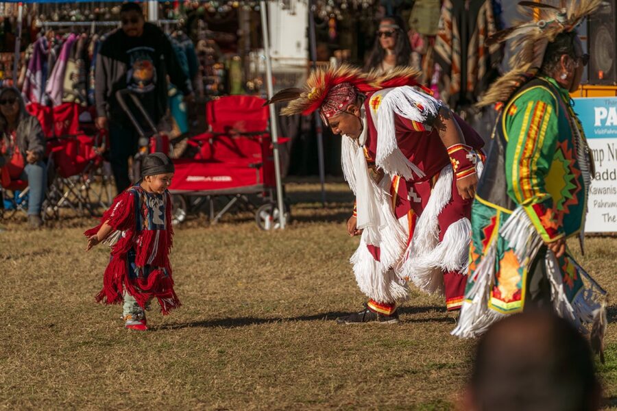 A young child and adult in traditional Native regalia dance together outdoors at a cultural event.