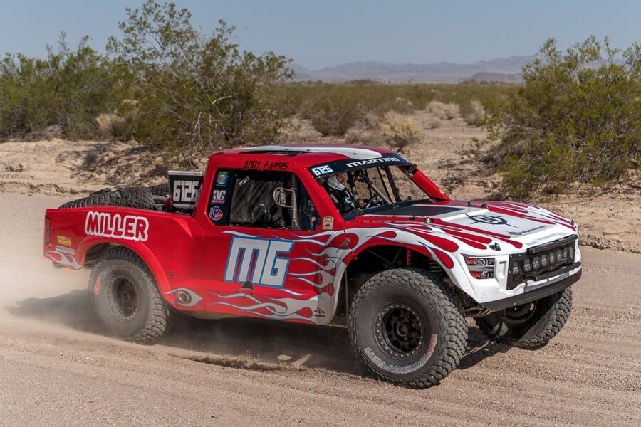 Red and white off-road race truck driving on a dirt track in a desert landscape with bushes and mountains.