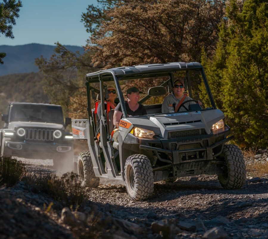 Three people drive a utility terrain vehicle on a rocky trail, with a jeep following behind in the background.