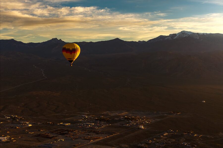 A colorful hot air balloon floats over a small town with mountains in the background at sunset.