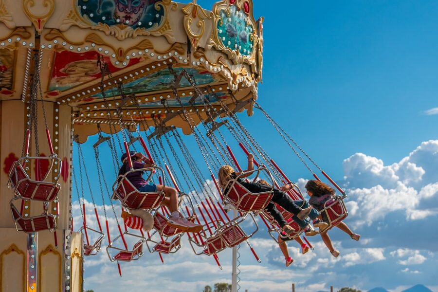 Children ride a colorful swing carousel at a fair under a bright blue sky with scattered clouds.