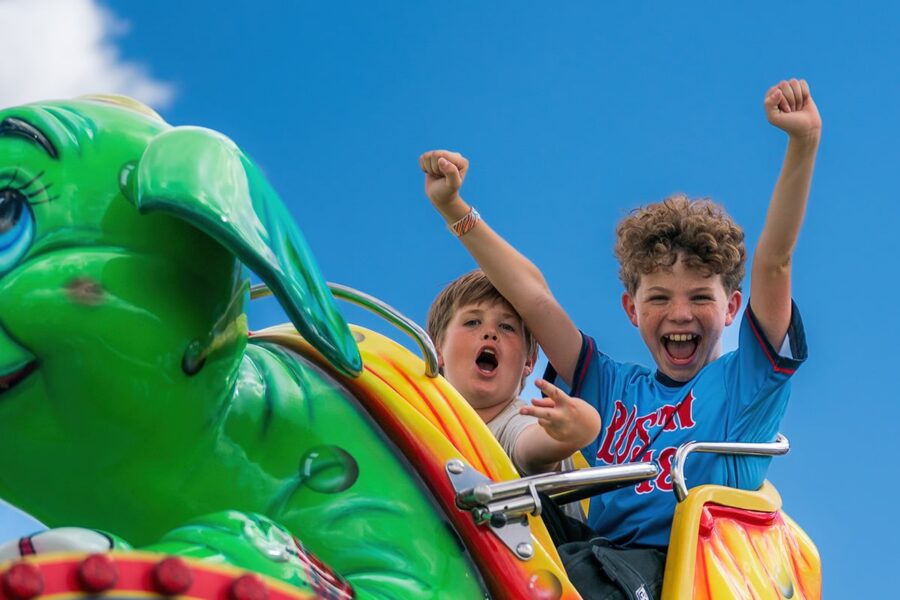 Two excited kids ride a green dragon amusement park ride, laughing and cheering under a bright blue sky.